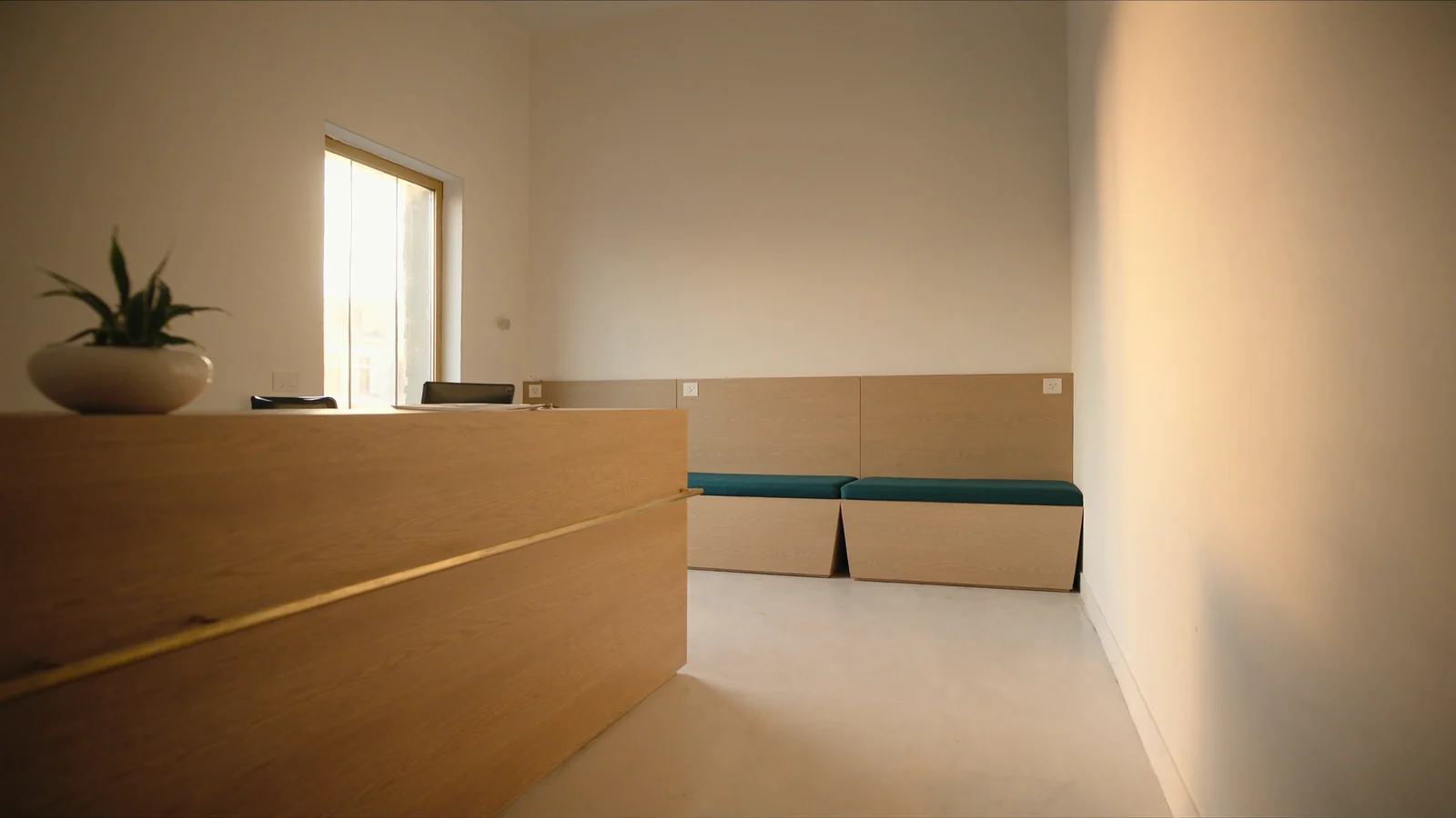 A newly finished medical spa reception, empty of people, with a pale oak desk, a built-in bench upholstered in teal, a potted plant on the counter and morning light from a tall window.