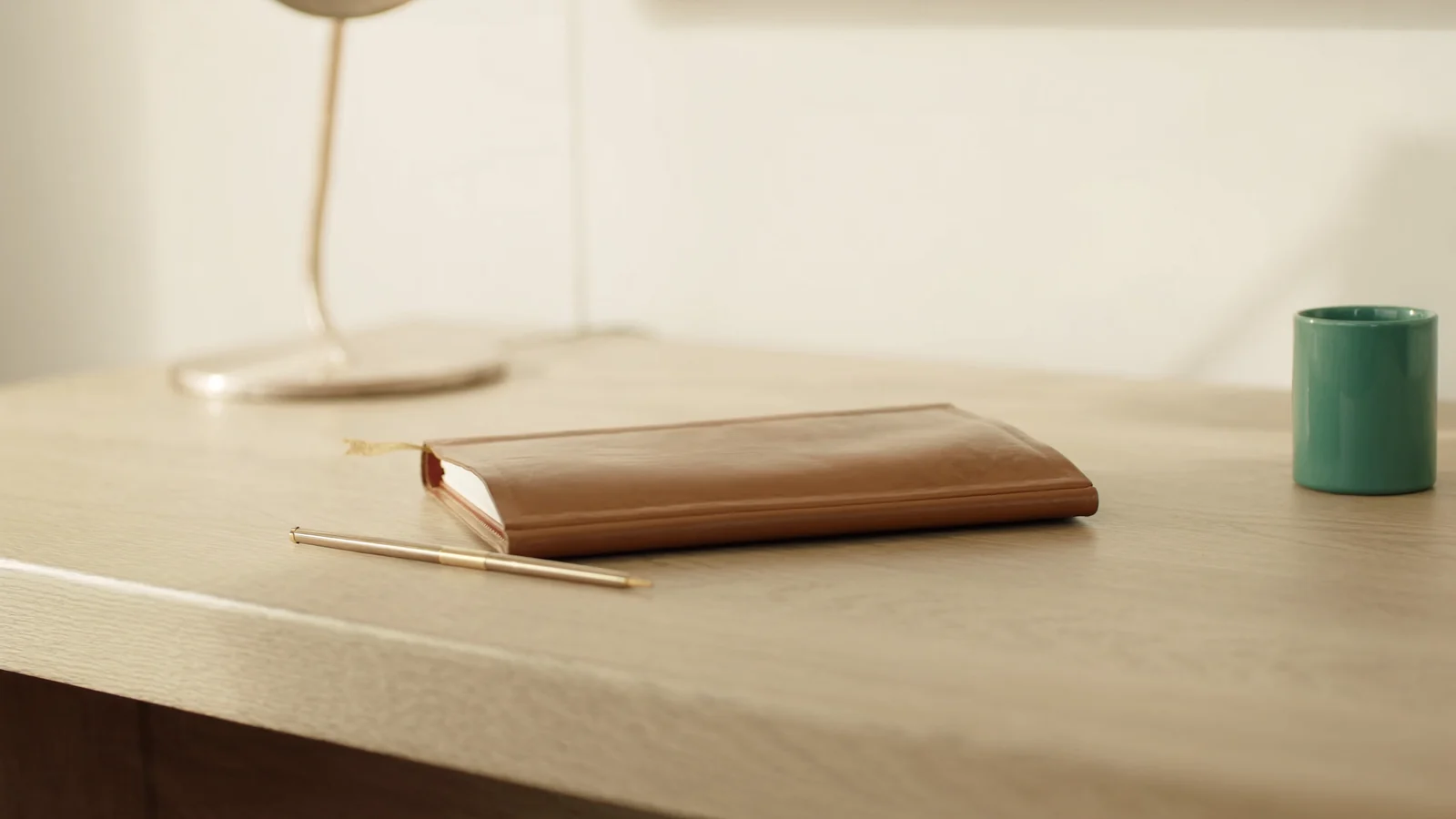 A plain tan leather notebook lying closed beside a slim brass pen on a pale oak desk, with a green-teal ceramic cup and an unlit brass lamp in soft morning light.