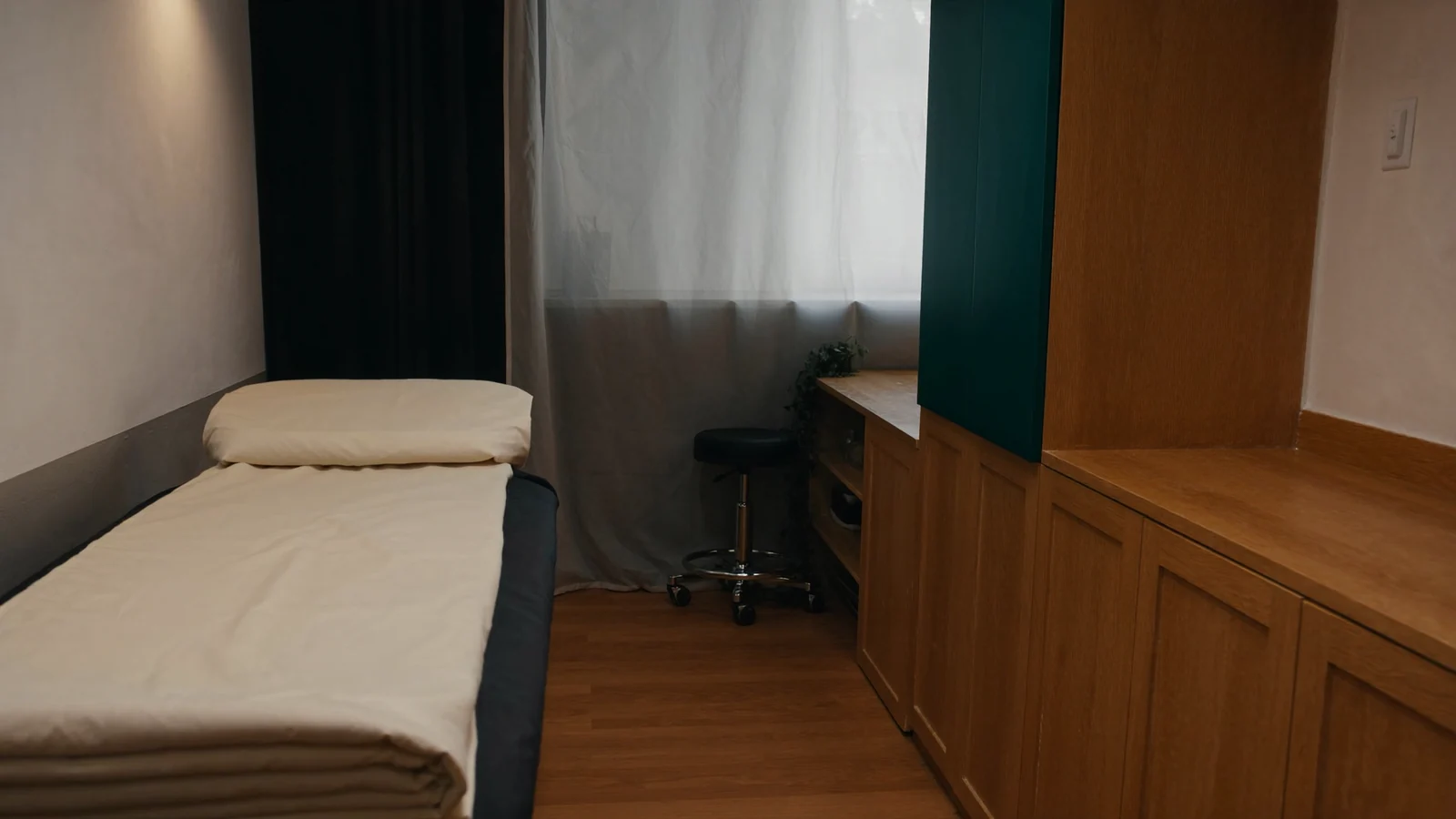 An empty, finished treatment room with honey-toned oak cabinetry, one cabinet front in deep teal, a bed made up with folded cream linen and a small rolling stool beside a curtained window.